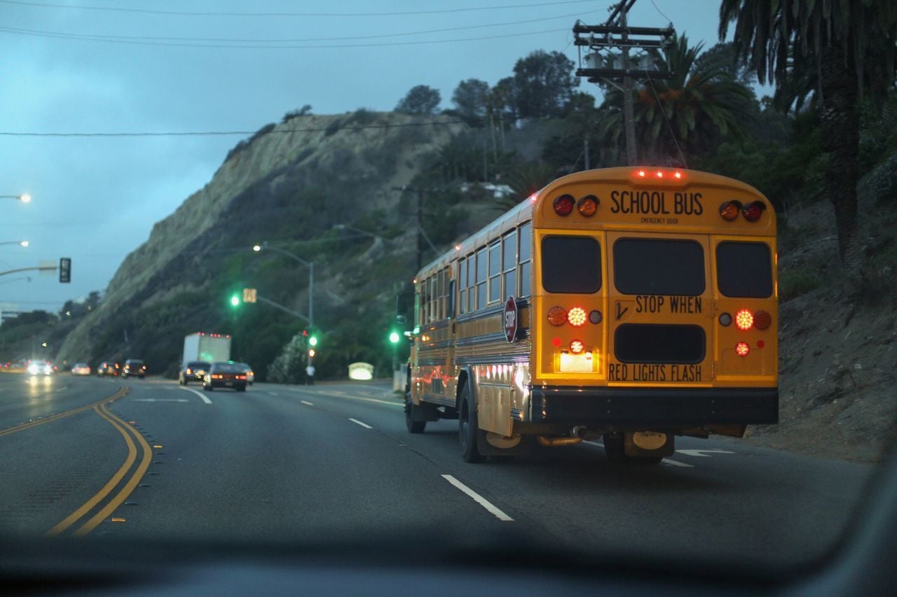 Yellow school bus driving on a coastal road near a cliff at dusk with red taillights lit.