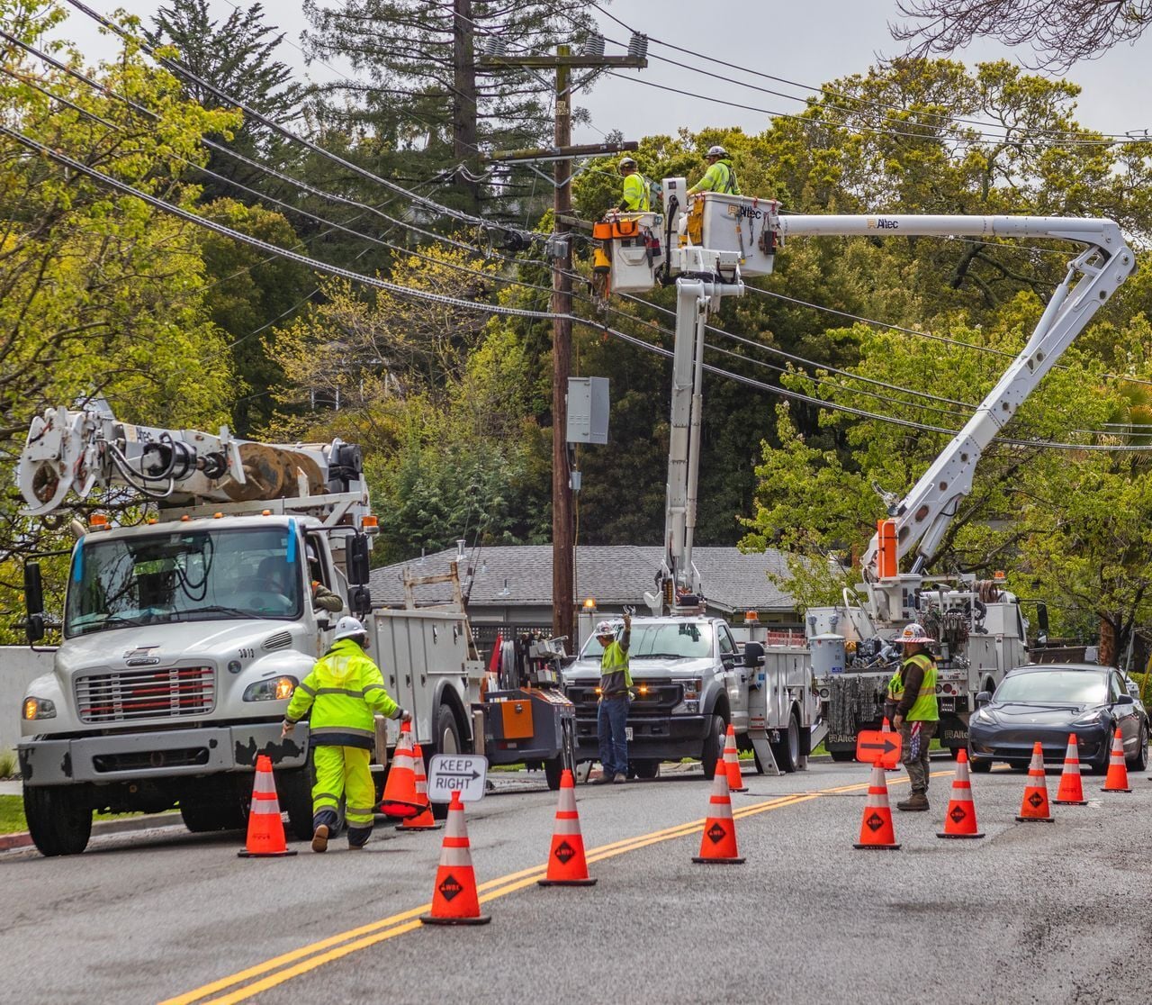Utility line workers in high-visibility gear work from bucket trucks to repair power lines on a residential street.